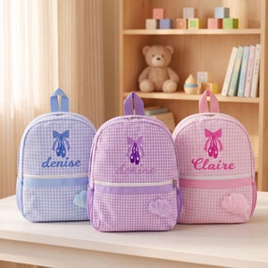 May include: Three backpacks in light blue, purple, and pink gingham patterns. Each features a top handle, a front zipper pocket, and a shell-shaped accent. Embroidered designs include ballet slippers with bows and the names Denise or Claire.