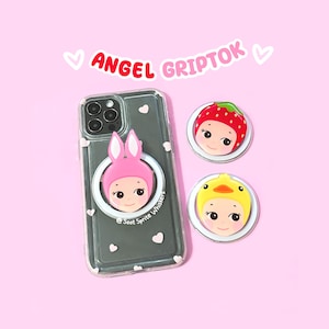 May include: A clear smartphone case with small white heart decals and a pink bunny-themed circular phone grip attached. Two additional circular phone grips are included: one featuring a strawberry design and one with a yellow duck design. The text ANGEL GRIPTOK and @ Soot Sprite Whiskers are visible.