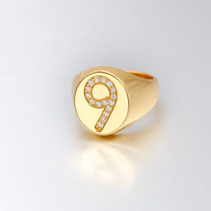 May include: A gold-toned signet ring featuring a circular face with the number nine embossed in the center. The number nine is outlined with small, clear, sparkling stones that contrast against the smooth, polished gold-colored metal surface.