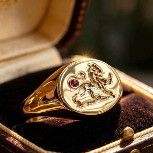 Könnte beinhalten: Ein polierter goldener Siegelring mit ovaler Platte, die ein eingraviertes Löwenmotiv zeigt, sowie einem kleinen, runden, tiefroten Edelstein, der nahe dem Löwenschwanz eingefasst ist. Der Ring hat eine glatte, reflektierende Oberfläche und eine konische Ringschiene.