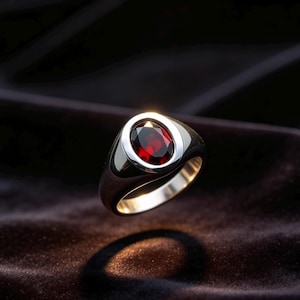 Könnte beinhalten: Ein Siegelring aus Silber mit einer polierten Metallschiene und einem zentralen, oval geschliffenen, tiefroten Granat-Edelstein. Der Ring hat eine glatte, reflektierende Oberfläche und eine Zargenfassung, die den facettierten Stein umschließt.