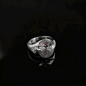 May include: A silver ring featuring a circular face with a relief design of two reaching hands. The background of the face has a sunburst pattern with small, embedded red stones scattered across the surface, creating a detailed metallic accessory.