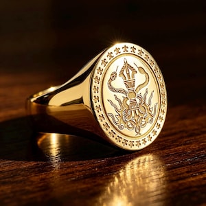 Pode incluir: Um anel de sinete em ouro polido com uma face oval apresentando um símbolo vajra gravado entrelaçado por duas serpentes. A borda da face do anel é decorada com um padrão repetitivo de flor-de-lis e a superfície metálica tem um acabamento brilhante.