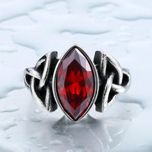 May include: A silver-toned ring featuring a central marquise-cut deep red gemstone. The band is crafted with intricate, raised knotwork detailing on both sides of the stone, creating a textured, metallic finish around the vibrant red center.
