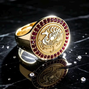 Op de afbeelding: Een gouden zegelring met een cirkelvormig vlak voorzien van een gegraveerd mythisch vogelontwerp. Het midden is omringd door een rand van kleine, ronde, dieprode robijnen. De ring heeft een gepolijste gouden band en een textuur op het embleem.