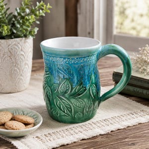 May include: A ceramic mug featuring a two-tone glaze with a deep green base and a vibrant blue upper section. The exterior is embossed with a repeating leaf pattern and a dotted texture near the rim. The interior of the mug is solid white.