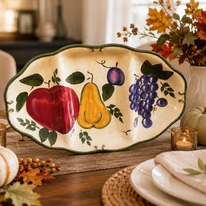 Puede incluir: Una fuente de cerámica ovalada con borde festoneado y base de color crema. La pieza presenta ilustraciones pintadas a mano de una manzana roja grande, una pera amarilla, una ciruela morada pequeña y un racimo de uvas moradas con hojas verdes.