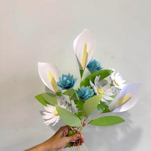 May include: A bouquet of paper flowers featuring white calla lilies with yellow centers, blue lotus flowers, and white daisies with yellow centers. The bouquet includes green leaves and is held together with wooden skewers.