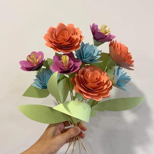 May include: A bouquet of paper flowers in shades of orange, purple, blue, and yellow. The flowers are arranged with green paper leaves and held together with wooden skewers. The bouquet is a handmade craft.