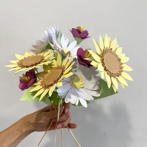 May include: A bouquet of paper flowers featuring sunflowers, daisies, and other blooms. The sunflowers have yellow petals and brown centers. The daisies are white and yellow. The bouquet is held together with wooden skewers and green paper.