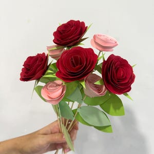 May include: A bouquet of paper roses in shades of red and pink, with green paper leaves. The flowers are held together with wooden skewers. The bouquet features several large red roses and smaller pink roses.