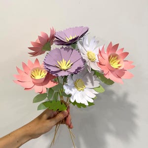 May include: A bouquet of handmade paper flowers in shades of pink, purple, and white, with yellow centers. The flowers are held together with wooden skewers and green paper leaves.
