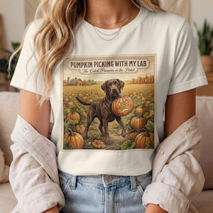 Könnte beinhalten: Ein weißes T-Shirt mit kurzen Ärmeln und einem Grafikdruck eines braunen Labrador-Hundes, der einen geschnitzten Kürbis in einem Kürbisfeld hält. Der Text PUMPKIN PICKING WITH MY LAB und The Cutest Pumpkin in the Patch ist abgebildet.