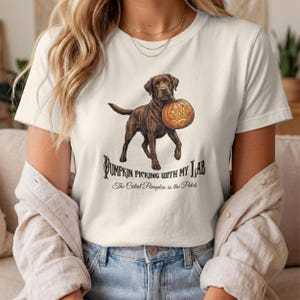 Könnte beinhalten: Ein cremefarbenes T-Shirt mit einer Grafik eines braunen Labrador Retrievers, der einen geschnitzten Kürbis im Maul hält. Der Text lautet: Pumpkin Picking With My Lab, The Cutest Pumpkin in the Patch, gedruckt in dunkelbrauner Schrift.