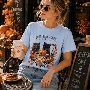 May include: A light blue t-shirt featuring a graphic of a black cat sitting at a table with a pumpkin-themed mug, a coffee cup, and a doughnut, surrounded by autumn leaves. The text reads PUMPKIN CAFE and Black Cat Cafe, with a small bird nearby.
