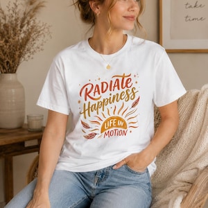 Puede incluir: Una camiseta blanca de manga corta con un diseño gráfico que incluye el texto "RADIATE Happiness" y "LIFE IN MOTION" en letras naranjas y rojas, acompañado de una ilustración de un sol, pequeñas estrellas y hojas de colores otoñales.