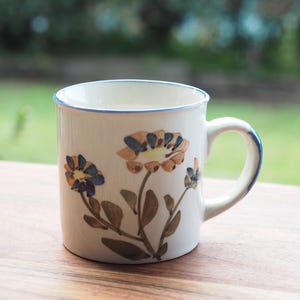 May include: A cream-coloured ceramic mug featuring a hand-painted floral design with brown stems, green leaves, and flowers in shades of blue and tan. The mug has a smooth cylindrical body, a curved handle, and a thin blue rim along the top edge.