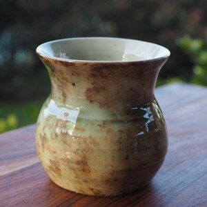 May include: A small ceramic vase with a rounded body and a flared rim. The surface features a glossy glaze with a mottled blend of pale green and earthy brown tones, creating a textured appearance across the exterior of the vessel.