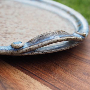 May include: A round ceramic serving tray with a speckled beige surface and a raised, blue-toned glazed rim. The tray features two integrated, curved handles on opposite sides, showcasing a mottled texture with brown and blue colour variations.