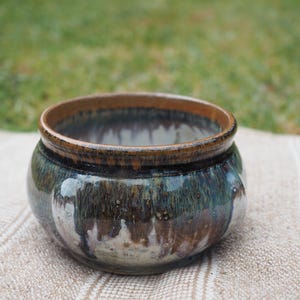 May include: A round ceramic bowl featuring a glossy glaze with blended tones of deep blue, forest green, and earthy brown. The rim is defined by a smooth, solid brown band, while the body displays a mottled, drip-like pattern of colours.