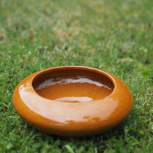 May include: A low-profile, circular ceramic bowl with a glossy, warm amber-orange glaze. The vessel features a wide, rounded rim that curves inward towards a smooth, concave centre, creating a simple, open-top design with a uniform finish.