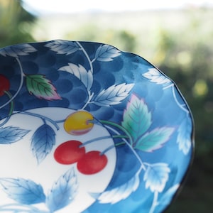 Puede incluir: Un cuenco de cerámica con borde festoneado que presenta un fondo azul intenso con un patrón repetitivo de hojas y tallos blancos. El diseño incluye cerezas de color rojo brillante y una cereza amarilla sobre un área central circular blanca.
