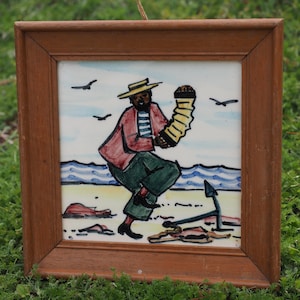 May include: A square ceramic tile framed in wood featuring a hand-painted illustration of a person wearing a straw hat, red jacket, and green trousers while playing an accordion on a beach with an anchor, blue waves, and flying birds.