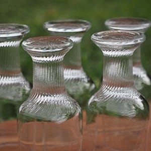 May include: A set of five clear glass tumblers positioned upside down. Each glass features a smooth, wide base and a neck with a distinct diagonal ribbed texture that transitions into a clear, smooth body. The glasses are arranged in a cluster.