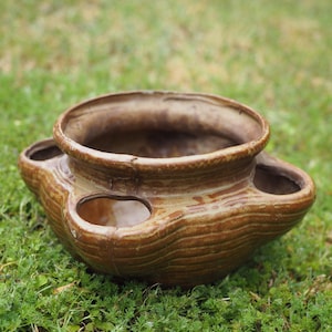 May include: A brown glazed ceramic strawberry planter with a wide central opening and five smaller circular side pockets. The pot features a textured, ribbed surface finish and a glossy, earthy brown glaze throughout the exterior structure.