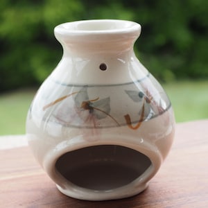 Może przedstawiać: Kremowy ceramiczny kominek do olejków o pękatym korpusie i szerokim, okrągłym otworze u podstawy. Zewnętrzna strona ozdobiona jest ręcznie malowanym motywem kwiatowym w stonowanych odcieniach niebieskiego i pomarańczowego, podkreślonym cienkimi szarymi liniami.