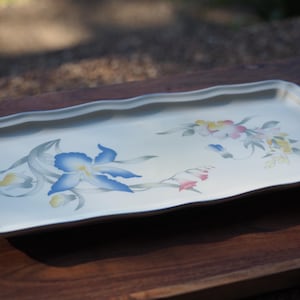 Op de afbeelding: Een rechthoekig, gebroken wit serveerblad met een geschulpte rand, voorzien van een geschilderd bloemmotief met een opvallende blauwe iris, kleinere gele knoppen en een tweede groep roze en gele bloemen met groen blad aan de rechterkant.