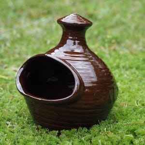 May include: A dark brown glazed ceramic salt pig with a glossy finish, featuring a wide, circular side opening for easy access and a small, pointed knob handle at the top. The exterior surface has subtle horizontal ridges around the body.