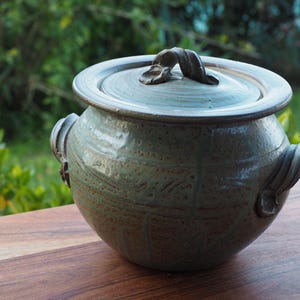May include: A round, sage green ceramic pot with a matching lid and two side handles. The surface features a subtle, grid-like texture with brown speckles throughout. The lid has a central, sculpted handle, and the overall finish is glossy.