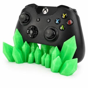 Peut inclure: Une manette de jeu vidéo sans fil noire reposant derrière un groupe de structures translucides en forme de cristaux pointus vert vif. La manette comprend une croix directionnelle, deux joysticks et quatre boutons d'action colorés marqués X, Y, A et B.