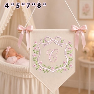 May include: A cream-colored fabric wall banner featuring a central embroidered letter C inside a shield-shaped frame, surrounded by pink flowers and green leaves. The banner hangs from a wooden rod with pink satin bows and text 4" 5" 7" 8".