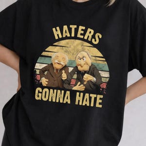 May include: A black short-sleeve t-shirt featuring a graphic print of two elderly puppet characters. Above and below the characters, the text reads "HATERS" and "GONNA HATE" in yellow. The background of the graphic is a sunset with horizontal stripes.