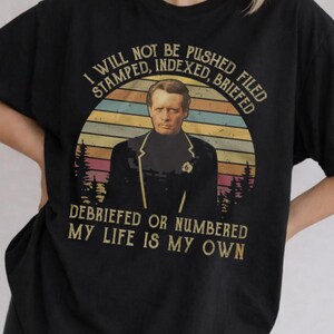 May include: A black short-sleeve t-shirt featuring a graphic of a man in a dark jacket. The text reads: I WILL NOT BE PUSHED FILED STAMPED, INDEXED, BRIEFED DEBRIEFED OR NUMBERED MY LIFE IS MY OWN. The design includes a sunset background.