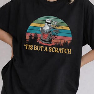 May include: A black t-shirt featuring a graphic of a knight in a helmet with a disintegrating arm. The design includes a sunset background with horizontal stripes in yellow, orange, and teal, and the text 'TIS BUT A SCRATCH' in yellow.