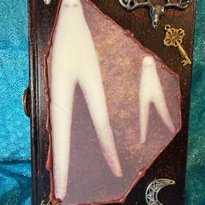 Può includere: Una copertina di libro scura e strutturata con un'immagine centrale di due figure bianche. L'immagine è incorniciata con un bordo rosso e adornata con ciondoli metallici, tra cui un cuore, una chiave, corna e una luna crescente. Il libro ha una chiusura.