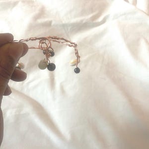 May include: A copper-colored wire bracelet featuring a twisted metal band design. The bracelet is adorned with several dangling charms, including small white seashells and smooth, round black beads attached to the wire with thin metal loops.