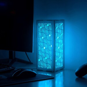 May include: A rectangular, illuminated lamp with a geometric pattern. The lamp emits a bright blue light, casting a cool glow on the surrounding desk and objects, including a computer monitor, mouse, and game controllers. The lamp's design features a series of interconnected shapes.