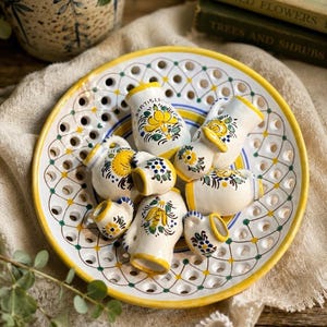 May include: A white ceramic bowl with a yellow rim and a lattice-style perforated border, containing a collection of small, hand-painted ceramic pitchers. Each pitcher features floral motifs in yellow, blue, and green, with one marked Bratislava.