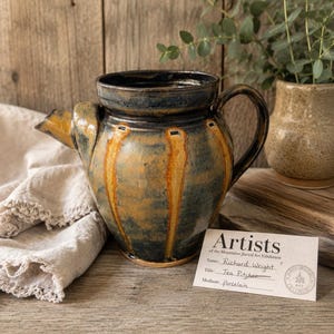 May include: A ceramic tea pitcher with a dark blue and amber glaze featuring vertical ribbed detailing, a curved handle, and a short spout. A small white exhibition card is placed in front, labeled Artists of the Mountains, Richard Wright, Tea Pitcher.