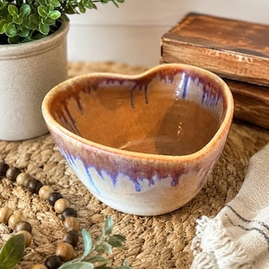 May include: A heart-shaped ceramic bowl with a glossy finish. The interior is a warm tan color, while the exterior features a reactive glaze with drips of deep purple and blue cascading down from the rim over a creamy off-white base color.