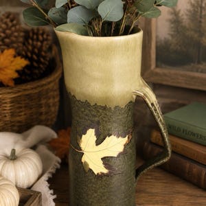 May include: A tall ceramic pitcher featuring a two-tone glaze with a light beige upper section and a dark forest green lower section. A single embossed maple leaf with yellow and brown tones is centered on the front, and a sturdy handle is attached.