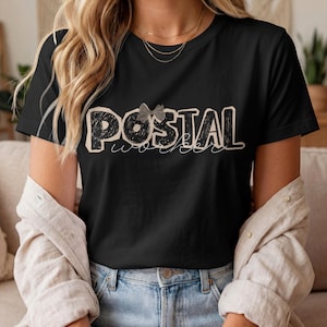 May include: A black short-sleeved t-shirt featuring the word POSTAL in large, distressed black block letters with a light-colored outline, and the word worker in a thin white cursive script below it, accented by a small gray bow graphic.