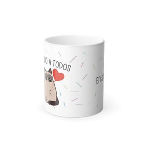 Puede incluir: Taza de cerámica blanca con un gato de dibujos animados con expresión de enfado y un corazón rojo. La taza tiene texto en español y líneas de colores. Un regalo humorístico para los amantes de los gatos.