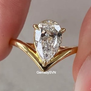 May include: A gold ring featuring a pear-shaped faceted clear gemstone set in a V-shaped band. The metal has a polished finish, and the stone is secured by small prongs. The text GemsbySVN is printed in small white letters at the base of the setting.