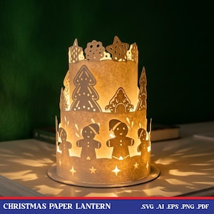 May include: A two-tiered cylindrical paper lantern featuring cut-out designs of gingerbread people, pine trees, stars, and decorative shapes. The lantern emits a warm, glowing light from within, casting intricate shadows on the surface below.