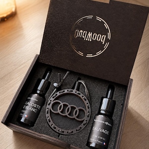 Op de afbeelding: Een donkerhouten geschenkdoos met twee glazen druppelflesjes van 10 ml met het label Lacoste L.12 Blanc en Sauvage, naast een ronde houten hanger met vier in elkaar grijpende ringen, alles geplaatst in een zwart schuimrubberen inzetstuk.